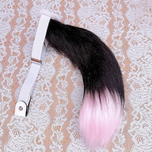 May include: A faux fur tail accessory featuring a long, curved design with black fur at the base transitioning into a light pink tip. The tail is attached to a white elastic belt with a metal adjustment buckle and a white snap-button closure.