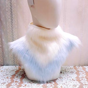 May include: A faux fur neck warmer featuring a gradient color pattern that transitions from a soft cream white at the top to a pale pastel blue at the bottom. The accessory has a thick, plush texture and is displayed on a mannequin neck form.