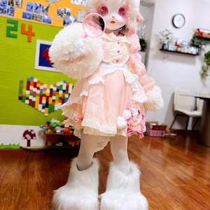 May include: A doll dressed in a pink and white outfit with a fluffy white headpiece and matching arm and leg coverings. The doll is holding a small, round mirror. The doll's dress has multiple layers of ruffles and lace.