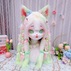 May include: A whimsical doll head with a white face, large green eyes, and red lips. The doll has white hair with green and pink streaks, styled in braids and adorned with bows and strawberry accents. The ears are cat-like, with pink and green details.