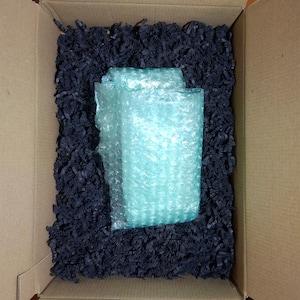 May include: A rectangular cardboard shipping box containing a central item wrapped in translucent light blue bubble wrap, surrounded by a thick layer of shredded black paper filler used for protective cushioning during transit and storage.