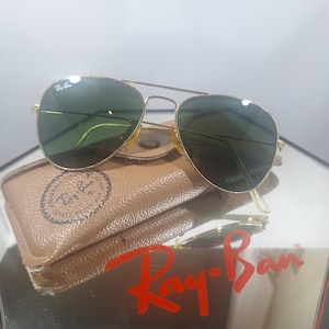 Peut inclure: Lunettes de soleil aviateur dorées avec des verres vert foncé et le logo Ray-Ban sur le coin supérieur. Les lunettes reposent sur un étui en cuir texturé beige avec un tampon Ray-Ban circulaire, au-dessus d'un grand logo Ray-Ban rouge sur un miroir.