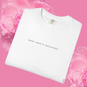 May include: White t-shirt with the text "smoke weed & masturbate" in a simple, sans-serif font. The shirt is displayed on a pink background with pink flowers in the corners. The shirt has a crew neck and short sleeves.
