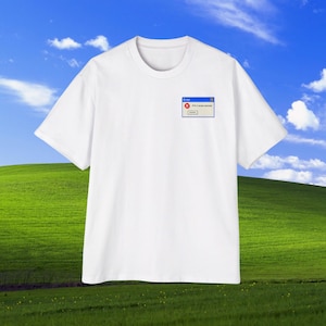 Könnte beinhalten: Weißes T-Shirt mit einer kleinen Grafik einer Computerfehlermeldung. Die Grafik enthält ein rotes "X" und das Wort "Error". Das Shirt befindet sich vor dem Hintergrund eines grünen Hügels und eines blauen Himmels mit Wolken.
