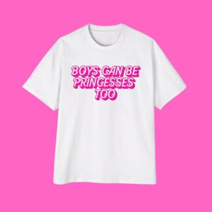 May include: White t-shirt with the text "BOYS CAN BE PRINCESSES TOO" in pink, outlined in a darker pink. The shirt is displayed against a bright pink background. The t-shirt has short sleeves and a crew neck.
