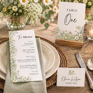 May include: A wedding stationery set featuring a menu card, a table number card labeled Table One, and a place card for Olivia Wilson. Each piece is decorated with a floral print of white daisies and blue wildflowers on white cardstock.