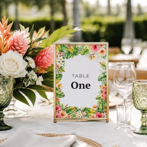 May include: A rectangular gold-framed table sign with the text "TABLE One" centered on a white background, surrounded by a colorful border of tropical flowers and green leaves, displayed upright on a table setting with glassware and floral decor.