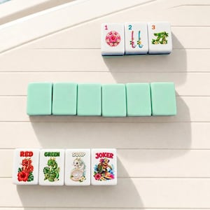 Pode incluir: Um conjunto de pe&ccedil;as retangulares de mahjong. A linha superior apresenta tr&ecirc;s pe&ccedil;as brancas com os n&uacute;meros 1, 2 e 3 e desenhos florais. A linha do meio tem seis pe&ccedil;as s&oacute;lidas em verde menta. A linha inferior mostra quatro pe&ccedil;as brancas com as etiquetas RED, GREEN, SOAP e JOKER.
