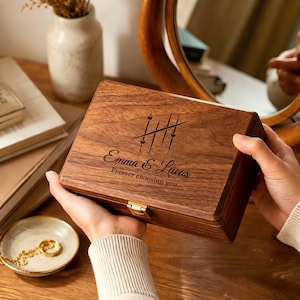 May include: A rectangular dark wood keepsake box with a gold-tone metal latch. The lid features a laser-engraved design of four vertical lines crossed by a diagonal line, small hearts, the names Emma & Lucas, and the text Forever choosing you.