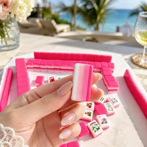 May include: A set of pink and white Mahjong tiles, with a focus on a single tile held in a hand. The tiles are arranged on a white surface, with some tiles displaying floral and animal designs. The background shows a tropical outdoor setting.