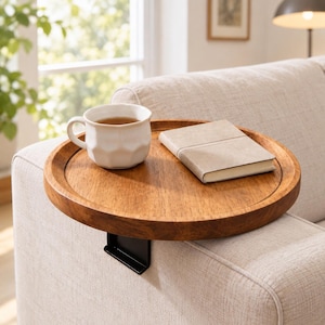 May include: A round wooden sofa arm tray with a natural brown finish and a black metal clip attachment. The tray holds a white ceramic mug with a faceted texture and a closed beige notebook, resting securely on the arm of a light-colored sofa.