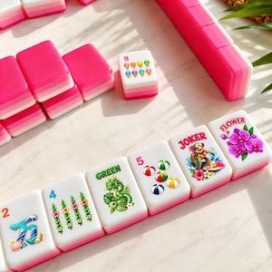 May include: A set of pink and white Mahjong tiles with various colorful illustrations and numbers. The tiles feature images of flowers, dragons, and ice cream cones. The set includes a pink tile holder.