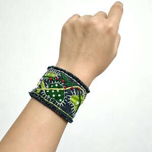 May include: A wide denim cuff bracelet featuring frayed edges and decorative embroidery. The design includes green fabric patches with white polka dots, accented by yellow stitched lines and small red circular embroidered details.