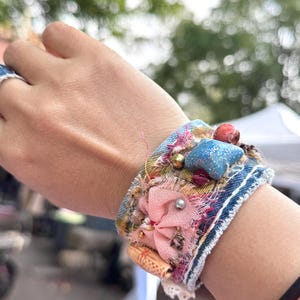 Puede incluir: Una pulsera de puño de mezclilla hecha a mano con un diseño de patchwork. La pulsera presenta una flor de tela rosa, adornos azules en forma de estrella y varias cuentas. El puño mide aproximadamente 5 cm de ancho.