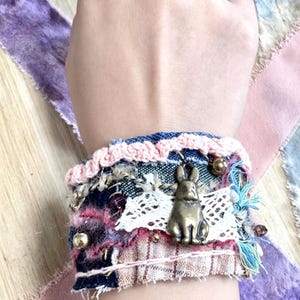 Pode incluir: Uma pulseira de tecido artesanal com um berloque de coelho de bronze. A pulseira apresenta camadas de ganga, renda e tecidos estampados em tons de rosa, azul e castanho. Acentuada com contas douradas e borlas azuis.