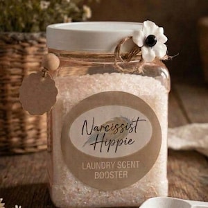 Peut inclure: Un bocal carré en plastique transparent rempli de cristaux blancs pour le linge. Le couvercle blanc est orné d'une fleur blanche et de ficelle. Une étiquette ronde indique Narcissist Hippie LAUNDRY SCENT BOOSTER. Une petite pelle blanche est posée.
