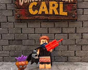 May include: A Lego minifigure with brown hair, a blue bandana, a black vest, and heart-patterned boxers, holding red dynamite. Beside it stands a brown cat figure wearing a purple crown and glasses, beneath a sign reading Dungeon Crawler Carl.