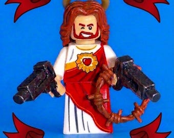 May include: A custom minifigure depicting a figure with long brown hair, a gold halo, and a white robe with a red sash featuring a heart emblem. The figure holds two black toy submachine guns and a brown barbed wire accessory against a blue background with red banners reading UZI JESUS and DUNGEON CRAWLER CARL.