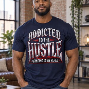 Può includere: Una t-shirt blu navy a maniche corte con un design grafico che riporta il testo ADDICTED TO THE HUSTLE in un carattere metallico argento e rosso, con il testo aggiuntivo GRINDING IS MY REHAB stampato in bianco sotto il design principale.