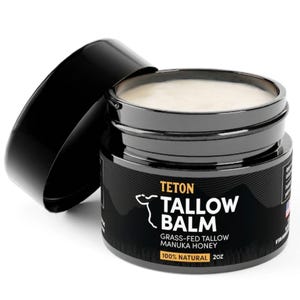Può includere: Un barattolo nero di Teton Tallow Balm con il coperchio aperto, che rivela un balsamo cremoso. L'etichetta presenta il testo "Tallow Balm" e "Grass-Fed Tallow Manuka Honey". Il barattolo è di 57 grammi.
