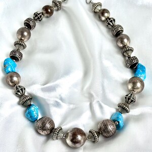 May include: A necklace featuring alternating turquoise stones and silver-toned beads. The silver beads include smooth spheres, ribbed spacers, and etched cylindrical components. The necklace is secured with a metal lobster claw clasp.
