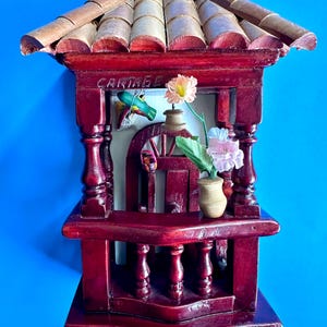 May include: A dark red wooden wall hanging shaped like a balcony with a tiled roof, featuring the word CARTAGO carved at the top. It includes a small vase with pink and white artificial flowers, a miniature bird figure, and two metal hooks at the base.