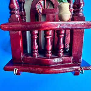 May include: A dark red stained wooden wall hanging featuring a miniature balcony with turned balusters, a small arched doorway, a tiny woven basket, and a small wooden vase containing artificial flowers, with two metal hooks at the base.