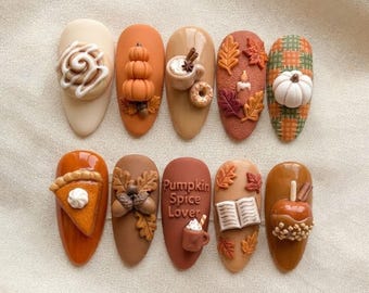 May include: A set of ten press-on nails featuring autumn-themed 3D decorations including a cinnamon roll, stacked pumpkins, a coffee mug, fall leaves, a candle, plaid patterns, pumpkin pie, acorns, a book, and a caramel apple with the text Pumpkin Spice Lover.