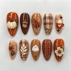 May include: A set of ten almond-shaped press-on nails featuring autumn-themed 3D designs. The collection includes motifs of pumpkins, acorns, fall leaves, plaid patterns, coffee mugs with whipped cream, cable knit textures, and floral accents.