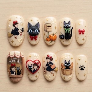 May include: A set of ten cream-colored press-on nails featuring illustrations of a girl with a red bow, a black cat, a bakery storefront with the text BAKERY, a mailbox, pumpkins, a red heart, and paw prints, accented with gold stars.
