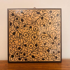 May include: A square wall plaque featuring a tan background with a black hand-drawn floral pattern of vines, leaves, and various flower shapes. The plaque has a dark brown border and a small metal hanging loop attached to the top center edge.