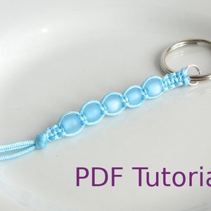 May include: A light blue beaded keyring with a silver key ring. The beads are strung on a light blue cord and tied with a knot at the end. The keyring is on a white surface. PDF Tutorial.