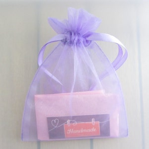 Puede incluir: Una peque&ntilde;a bolsa de regalo transparente de color lavanda con un cord&oacute;n de cinta de raso. En el interior, se ve un paquete rosa, con una peque&ntilde;a etiqueta que dice "Handmade" en un rect&aacute;ngulo rojo. La bolsa est&aacute; atada en la parte superior.