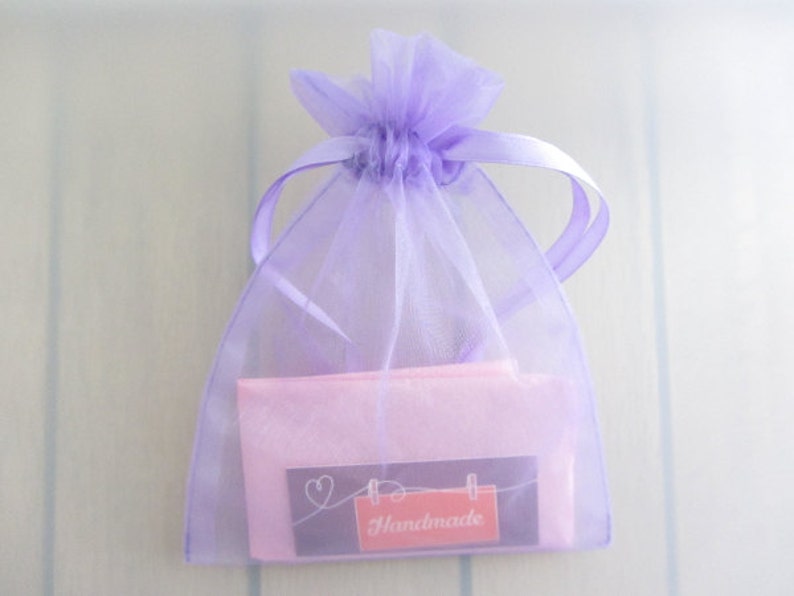 Puede incluir: Una peque&ntilde;a bolsa de regalo transparente de color lavanda con un cord&oacute;n de cinta de raso. En el interior, se ve un paquete rosa, con una peque&ntilde;a etiqueta que dice "Handmade" en un rect&aacute;ngulo rojo. La bolsa est&aacute; atada en la parte superior.