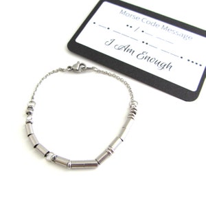 May include: A silver bracelet with a chain and small cylindrical beads. The bracelet is accompanied by a black card with the text "Morse Code Message" and the message "I Am Enough" written in Morse code.