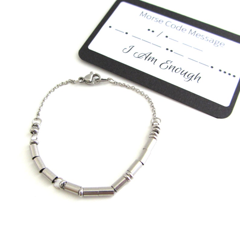 Stainless steel I am enough Morse Code bracelet (thin round and cylinder beads spell out the message) with a fine cable chain and lobster claw clasp. The bracelet lays next to a card that has printed on it the I am enough Morse Code Message.