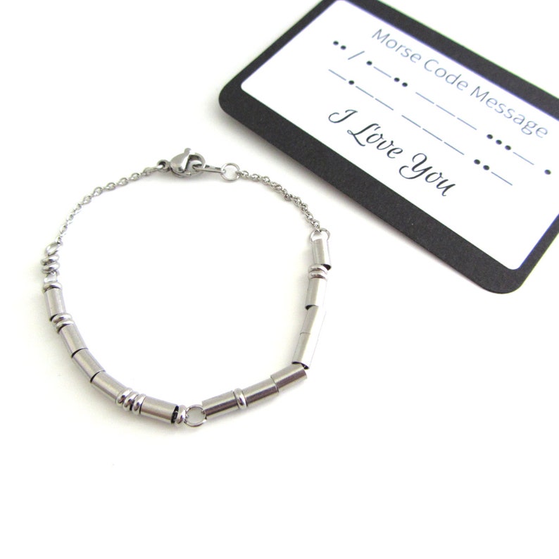 Stainless steel I love you Morse Code bracelet (thin round and cylinder beads spell out the message) with a fine cable chain and lobster claw clasp. The bracelet lays next to a card that has printed on it the I love you Morse Code Message.