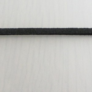 Puede incluir: Primer plano de un fino cord&oacute;n de ante negro. El cord&oacute;n est&aacute; centrado y se extiende horizontalmente por el marco. La textura del cord&oacute;n parece ligeramente &aacute;spera. El fondo es una superficie lisa de color blanquecino.