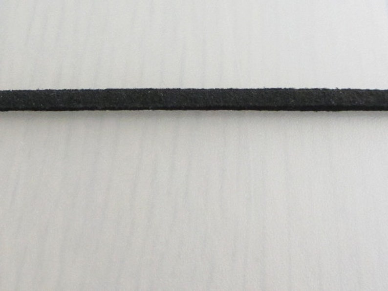 Puede incluir: Primer plano de un fino cord&oacute;n de ante negro. El cord&oacute;n est&aacute; centrado y se extiende horizontalmente por el marco. La textura del cord&oacute;n parece ligeramente &aacute;spera. El fondo es una superficie lisa de color blanquecino.