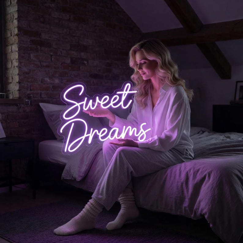 Może przedstawiać: Neonowy znak z napisem Sweet Dreams wykonanym kursywą. Znak emituje jasne, fioletowe światło, kt&oacute;re delikatnie oświetla otoczenie. Tekst jest wyraźnie czytelny, a słowo Sweet znajduje się bezpośrednio nad słowem Dreams.