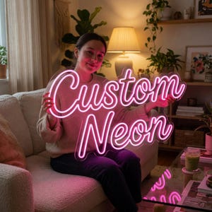 Może przedstawiać: Spersonalizowany neon z napisem Custom Neon wykonanym jasnor&oacute;żową kursywą. Znak jest zamontowany na przezroczystym akrylowym podkładzie i emituje żywą r&oacute;żową poświatę, wyraźnie prezentując tekst w dw&oacute;ch liniach na neutralnym tle.