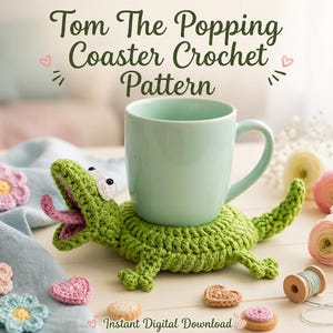 May include: A green crochet coaster shaped like a lizard with an open mouth and a light blue mug resting on its back. The text reads Tom The Popping Coaster Crochet Pattern and Instant Digital Download. Small crochet flowers and hearts are nearby.