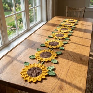 May include: A long table runner composed of seven connected crochet sunflower motifs. Each sunflower features a dark brown circular center, bright yellow petals, and small green leaves attached to the sides, arranged in a linear pattern.