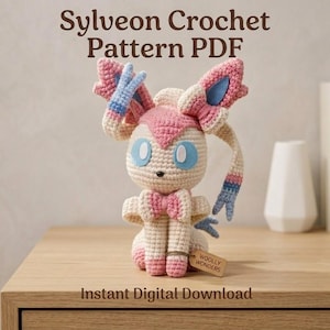 Puede incluir: Muñeca Sylveon de ganchillo con detalles rosas, blancos y azules. La muñeca tiene orejas grandes, un lazo y una etiqueta que dice "WOOLLY WONDERS". También se ve el texto "Sylveon Crochet Pattern PDF" y "Instant Digital Download".