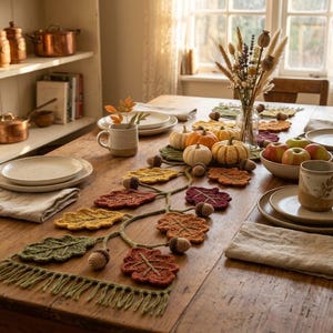Pode incluir: Um caminho de mesa de crochê com um design de videira com várias folhas em cores de outono em tons de verde, amarelo, laranja e vermelho, acentuado com pequenas bolotas de crochê e finalizado com uma borda de franjas na extremidade.