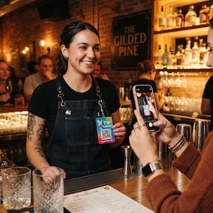 May include: A rectangular plastic tip card with a colourful gradient background, featuring a cocktail illustration, a QR code, and the text TIP SARAH SCAN TO TIP. The card is held up in a bar setting, displaying a clear, readable design for digital payments.