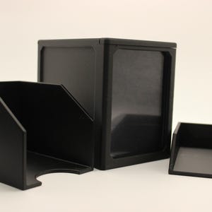May include: A matte black storage system consisting of a tall rectangular box with a front-facing window, a medium-sized open-front bin with a curved cutout at the base, and a small shallow tray, all featuring a smooth, uniform finish.