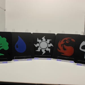 May include: Five square black coasters, each featuring a distinct colored icon: a green tree, a blue water droplet, a white sun, a red flame, and a white skull. Each coaster is displayed upright on a small, clear plastic stand for visibility.