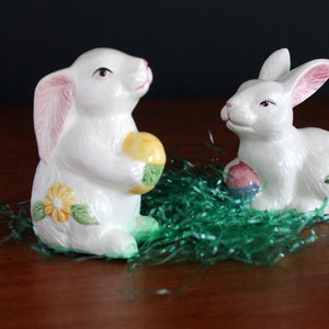 May include: Two white ceramic bunny figurines with pink-lined ears and floral accents. One bunny holds a yellow and green egg, the other a pink and blue egg. They sit on green decorative grass, perfect for Easter.