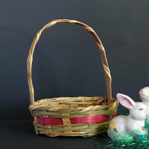 May include: A woven basket with a handle, featuring green and red accents, sits next to two white ceramic bunny figurines with pink ears. The bunnies are on a bed of green decorative grass, suggesting a springtime or Easter theme.